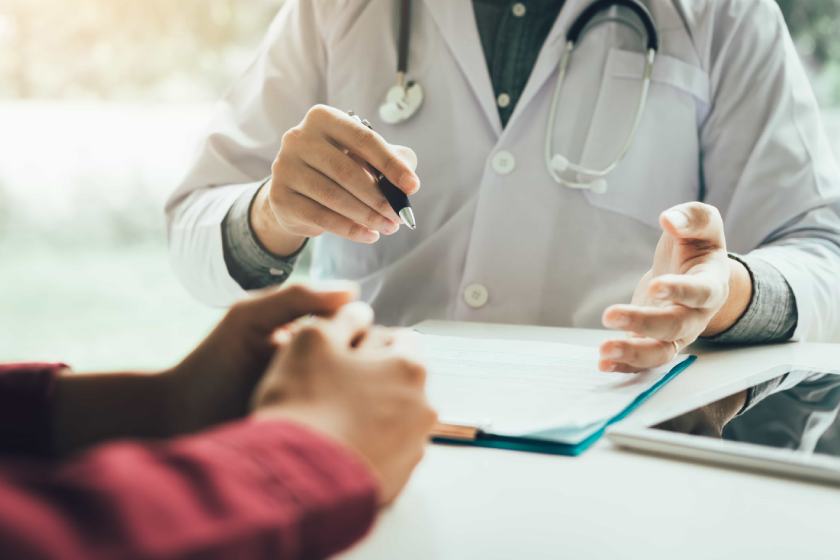 Medico in camice bianco discute un referto con un paziente, mostrando un documento su una cartella. 