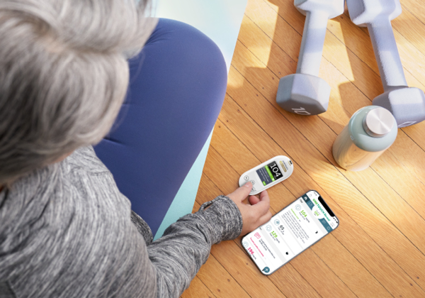 An older adult sitting on a yoga mat, checking their glucose level with a OneTouch Verio Reflect meter while a smartphone displays health data, alongside dumbbells and a water bottle.