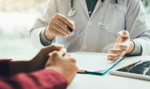 Medico in camice bianco discute un referto con un paziente, mostrando un documento su una cartella. 